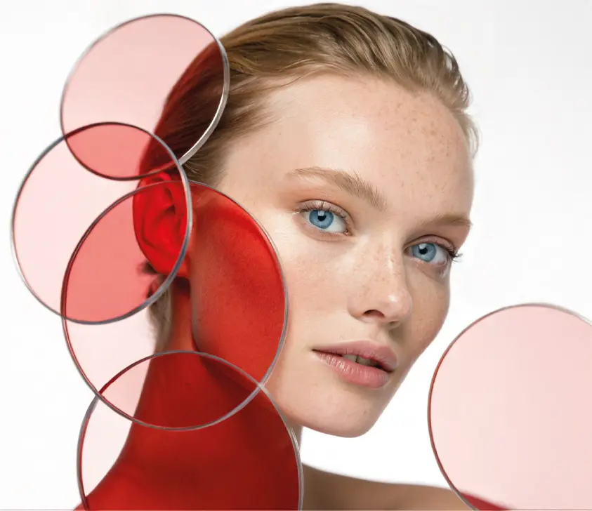A woman with blue eyes and light skin poses with her face framed by several translucent red and pink circular discs against a white background. The circles create layered color effects on her face.
