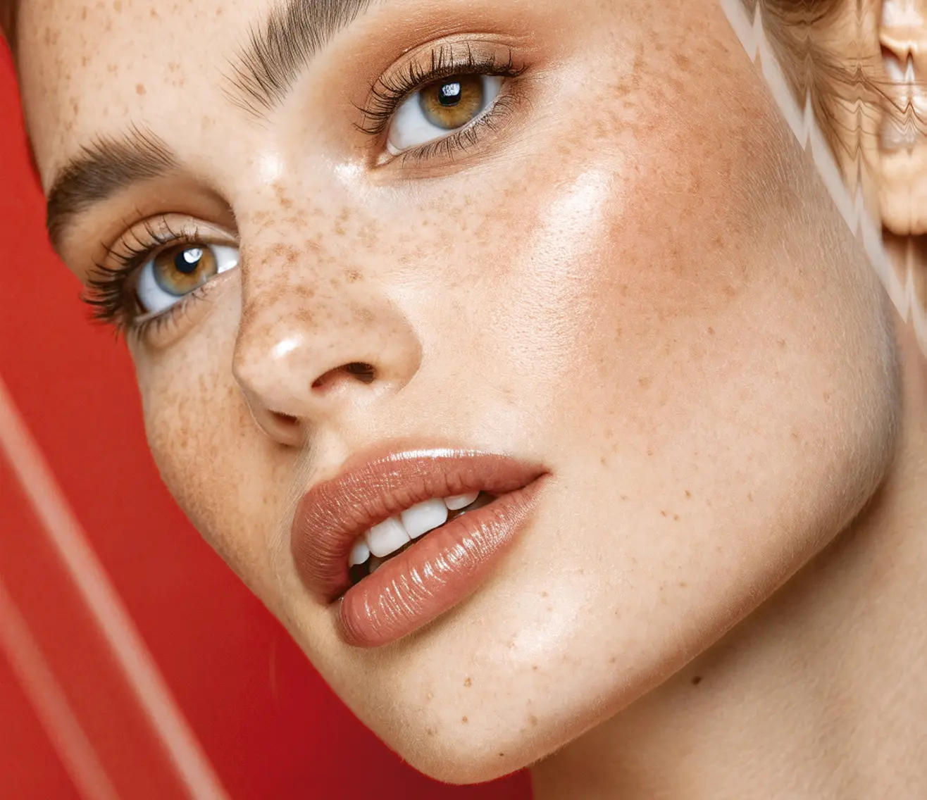 A close-up of a woman's face with glowing skin, freckles, full lips, and green eyes, set against a red background with white diagonal lines.