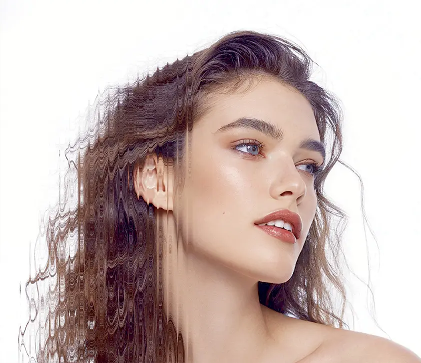 A woman with long wavy brown hair and natural makeup looks to the side. The left half of her hair appears digitally distorted, creating a rippled effect. The background is white.