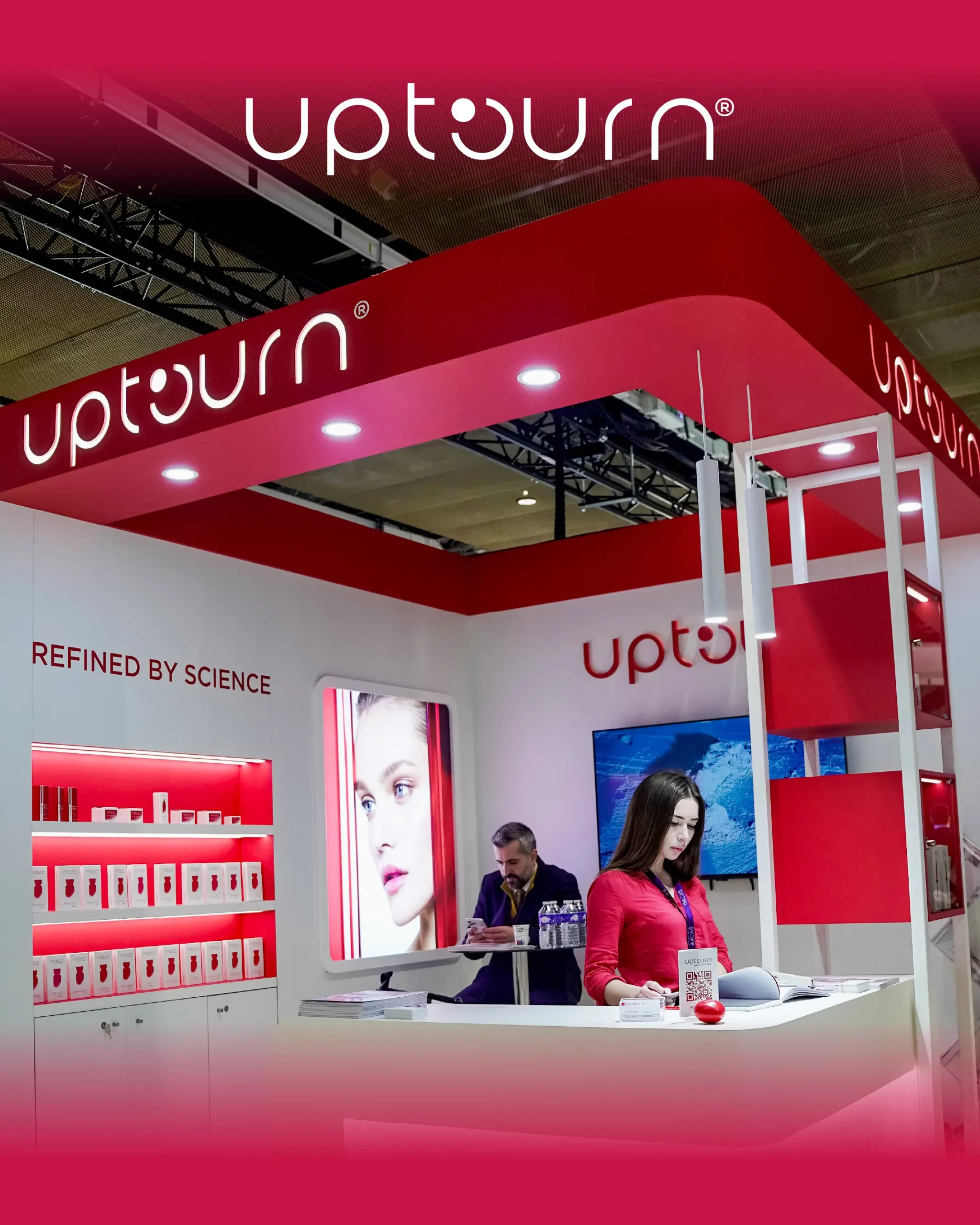 A woman stands at a modern, red-and-white “Uptourn” exhibition booth with shelves of products, a large model photo, and the slogan “Refined by Science” on the wall. Another person is seated in the background.