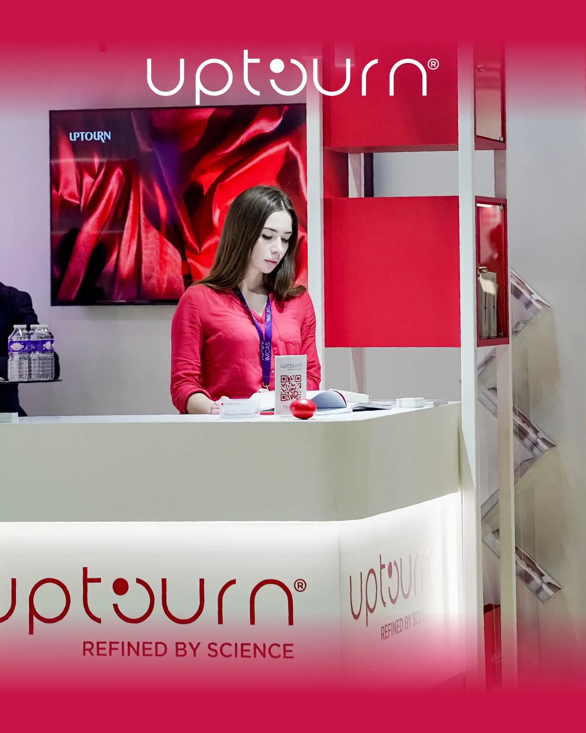 A woman in a red shirt stands behind a white counter at an Uptourn booth, with red and white branding and a TV displaying a red background behind her. The slogan "Refined by Science" is visible on the counter.