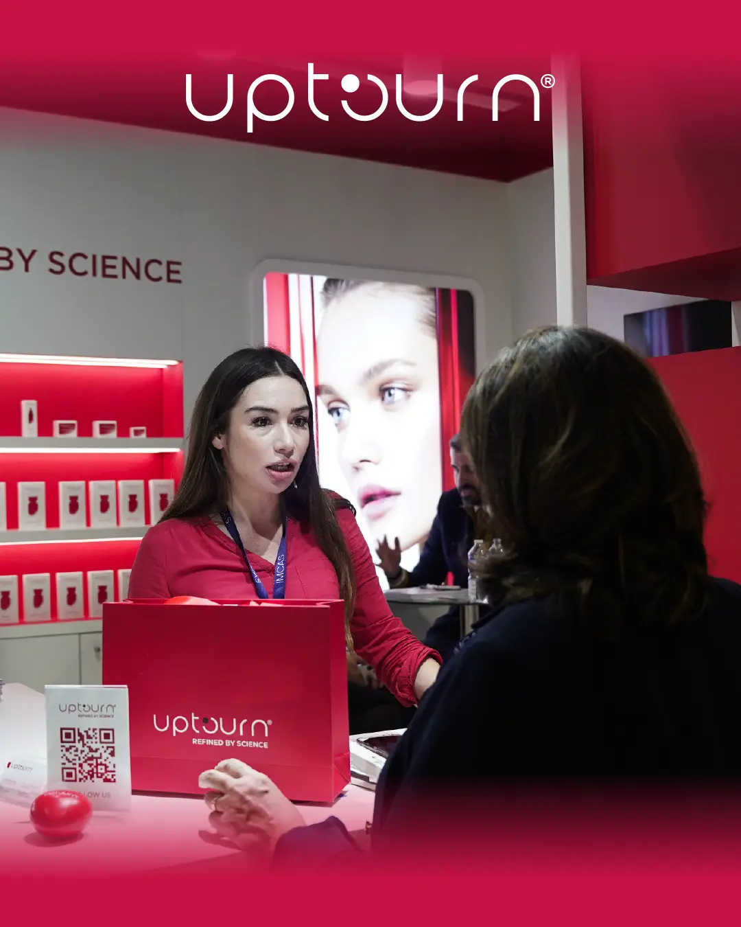 A woman at an "Upturn" booth speaks to a visitor, displaying a branded box and QR code. The backdrop shows shelves with products and a large image of a woman's face. The scene has a modern, pink-themed design.