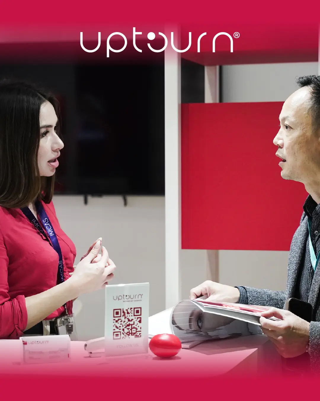 A woman and a man have a conversation at an Uptourn booth. The woman stands near a table with Uptourn brochures and a QR code, while the man holds papers. The scene has a red and white color theme.