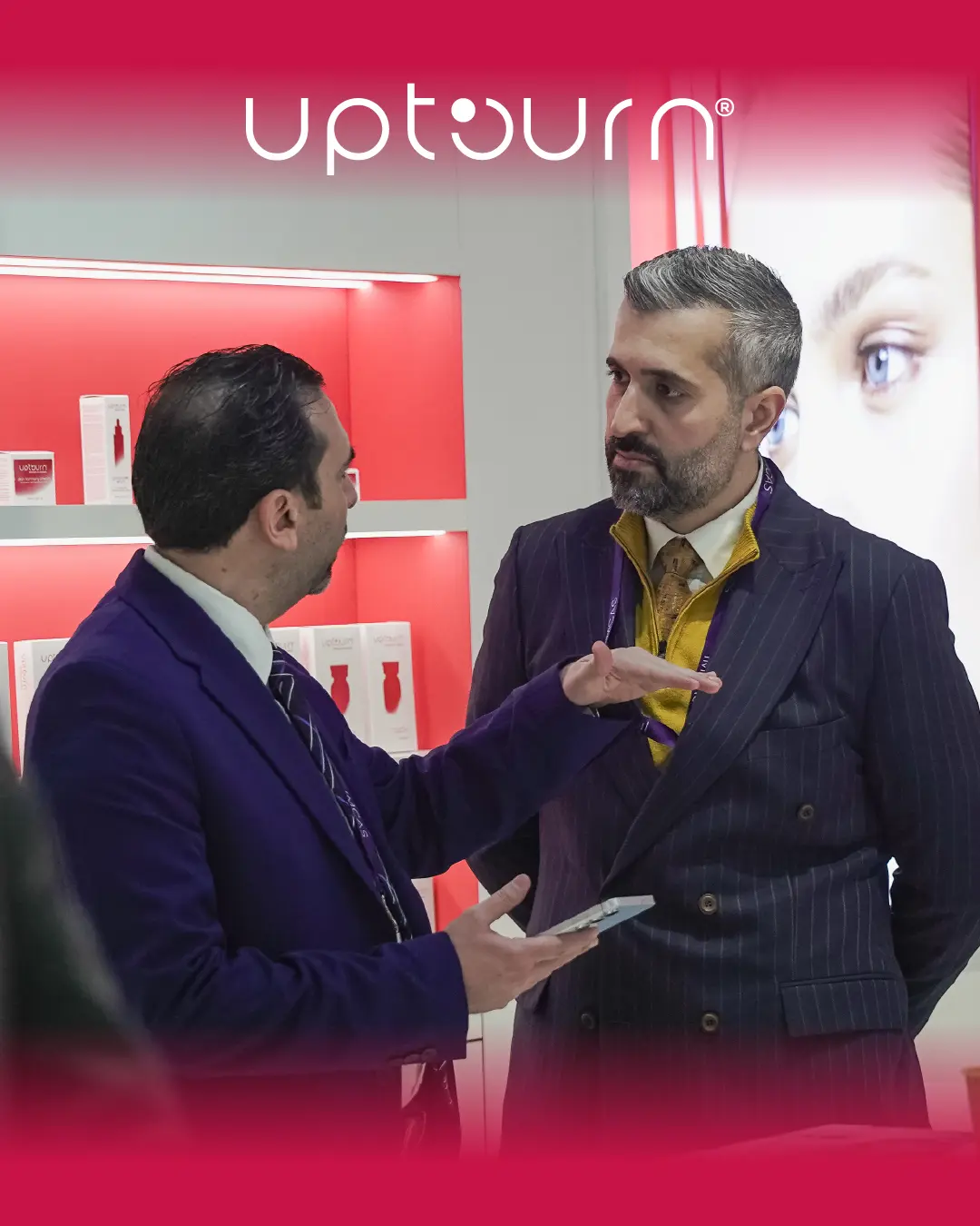 Two men in suits have a serious conversation indoors, surrounded by red and white product displays, with the word "upturn" at the top of the image and a blurred close-up face in the background.