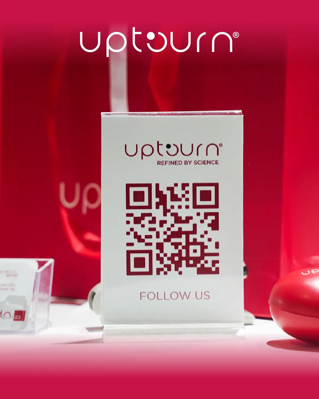 A tabletop display with a QR code and the text “upturn® REFINED BY SCIENCE” and “FOLLOW US,” surrounded by red upturn-branded objects and bags.