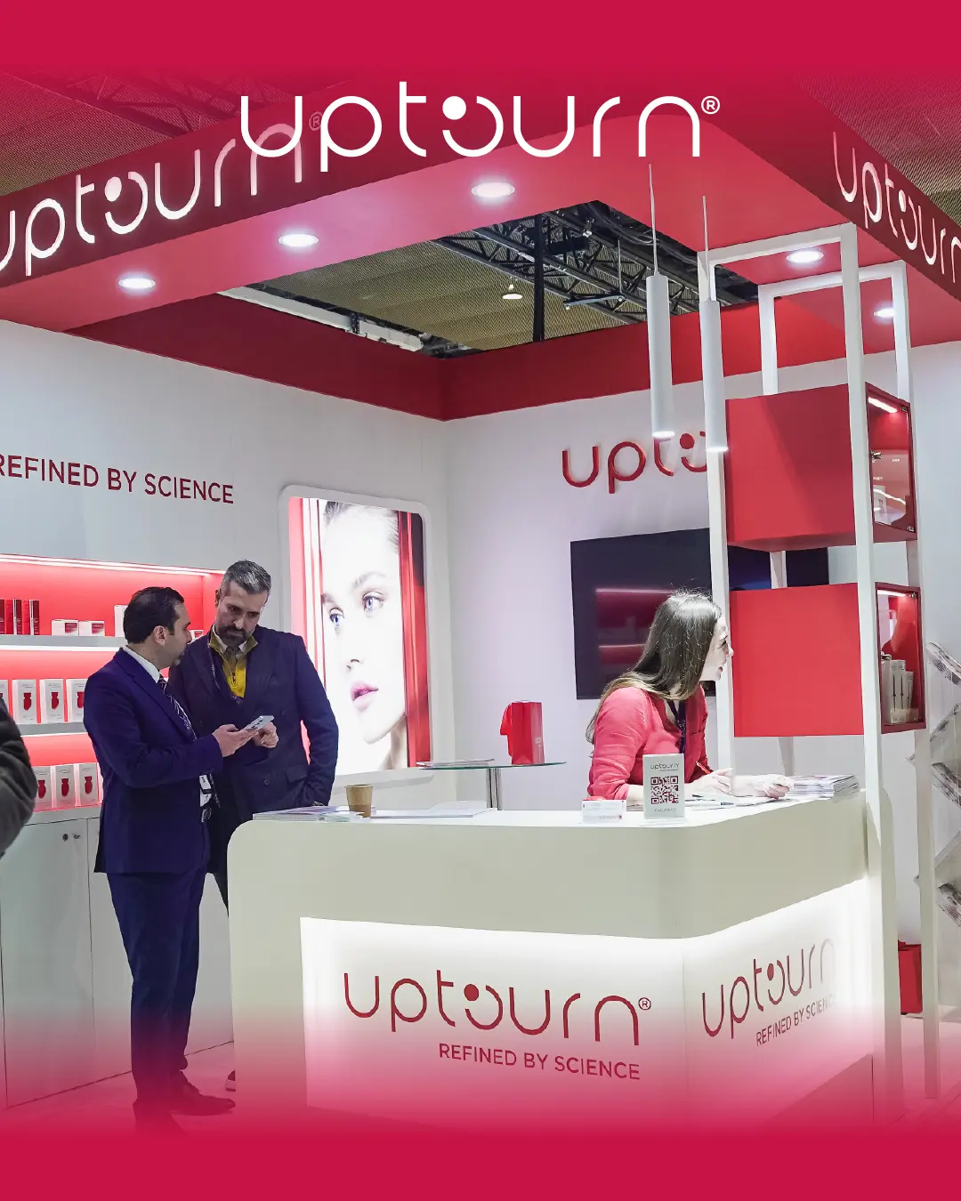 Three people interact at a modern, brightly lit trade show booth for "Upturn," a skincare brand. The booth features red and white decor, product displays, branding, and a large image of a woman's face.